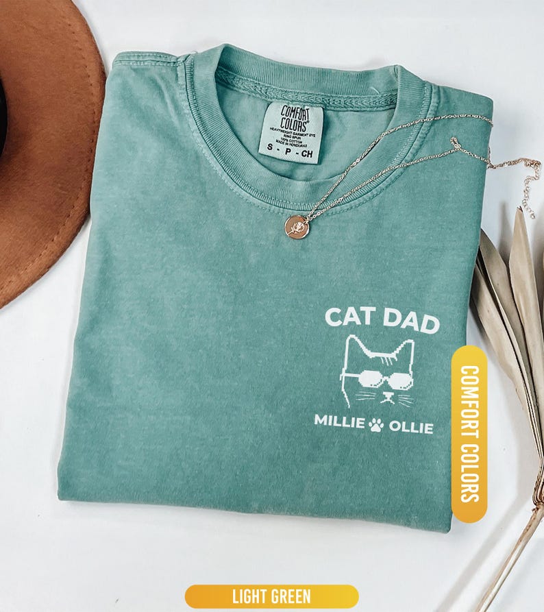 May include: Light green t-shirt with the text "CAT DAD" and a cat graphic wearing sunglasses. The shirt also features the names "Millie" and "Ollie" below the cat graphic. A gold necklace and a brown hat are also visible.