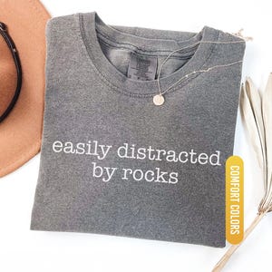 May include: A gray Comfort Colors t-shirt with the phrase "easily distracted by rocks" in white lettering. A gold necklace with a circular pendant rests on the shirt. A brown hat and dried plant are in the background.