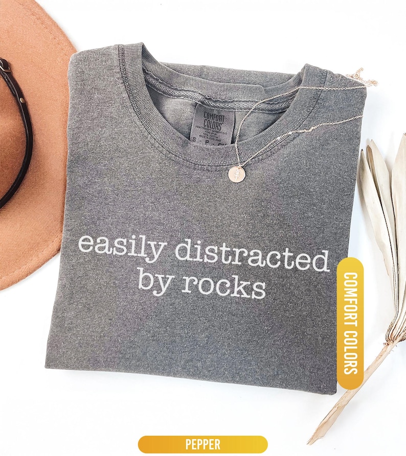 May include: A gray Comfort Colors t-shirt with the phrase "easily distracted by rocks" in white lettering. A gold necklace with a circular pendant rests on the shirt. A brown hat and dried plant are in the background.