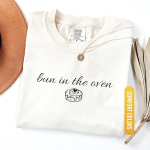 May include: Ivory-colored t-shirt with the phrase "bun in the oven" and a graphic of a cinnamon roll. The shirt has a crew neck and short sleeves. A gold necklace with a pendant is draped on the shirt. The shirt is labeled "Comfort Colors".