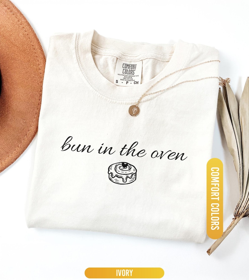 May include: Ivory-colored t-shirt with the phrase "bun in the oven" and a graphic of a cinnamon roll. The shirt has a crew neck and short sleeves. A gold necklace with a pendant is draped on the shirt. The shirt is labeled "Comfort Colors".