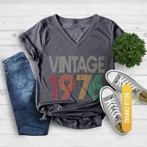 May include: Dark gray heather V-neck t-shirt with the text "VINTAGE 1976" in a retro design. The shirt is paired with blue jeans and gray canvas sneakers. The shirt is made of Bella Canvas material.