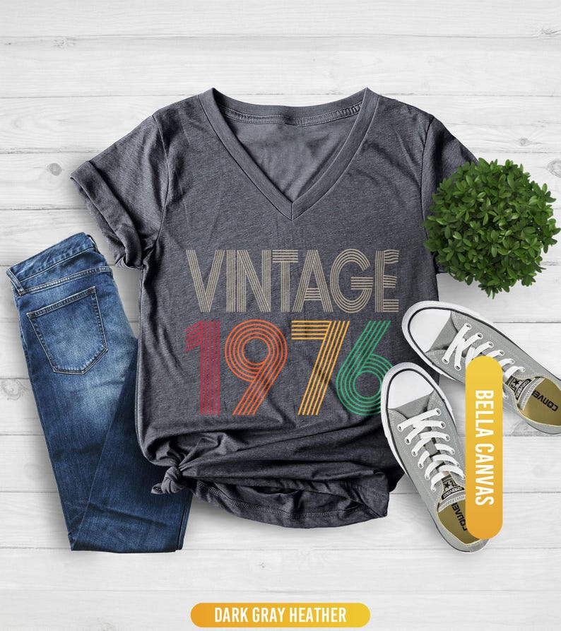 May include: Dark gray heather V-neck t-shirt with the text "VINTAGE 1976" in a retro design. The shirt is paired with blue jeans and gray canvas sneakers. The shirt is made of Bella Canvas material.