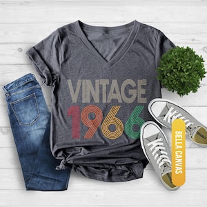 Pu&ograve; includere: T-shirt girocollo grigio scuro melange con la scritta "VINTAGE 1966" in un design retr&ograve;. La maglietta &egrave; abbinata a jeans blu e scarpe da ginnastica grigie. Le parole "BELLA CANVAS" sono stampate su un'etichetta gialla.