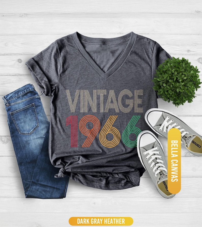 Pu&ograve; includere: T-shirt girocollo grigio scuro melange con la scritta "VINTAGE 1966" in un design retr&ograve;. La maglietta &egrave; abbinata a jeans blu e scarpe da ginnastica grigie. Le parole "BELLA CANVAS" sono stampate su un'etichetta gialla.