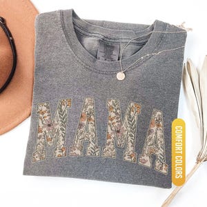 Op de afbeelding: Een grijs Comfort Colors t-shirt met het woord "MAMA" in bloemenprint. Het shirt is opgevouwen en heeft een ketting met een hanger. Een bruine hoed en een gedroogde plant staan op de achtergrond. Het shirt is gelabeld "Pepper".