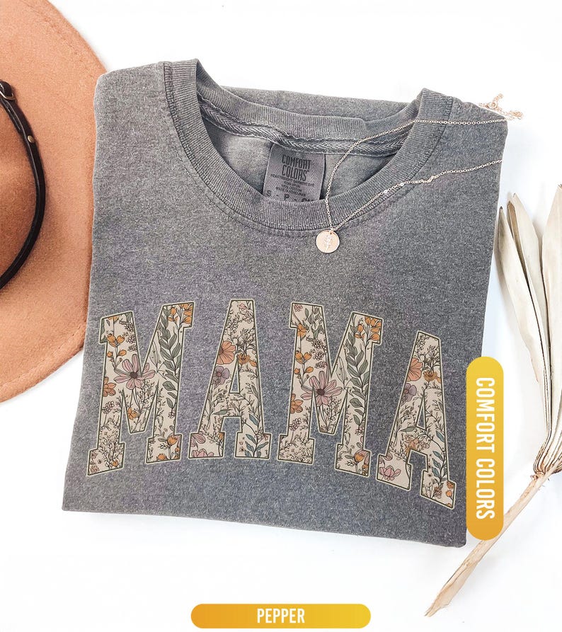 Op de afbeelding: Een grijs Comfort Colors t-shirt met het woord "MAMA" in bloemenprint. Het shirt is opgevouwen en heeft een ketting met een hanger. Een bruine hoed en een gedroogde plant staan op de achtergrond. Het shirt is gelabeld "Pepper".