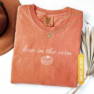 May include: A coral-colored t-shirt with the phrase "bun in the oven" printed in white, along with a graphic of a cinnamon roll. The shirt has a crew neck and a small tag that says "Comfort Colors". A gold necklace and a brown hat are also visible.