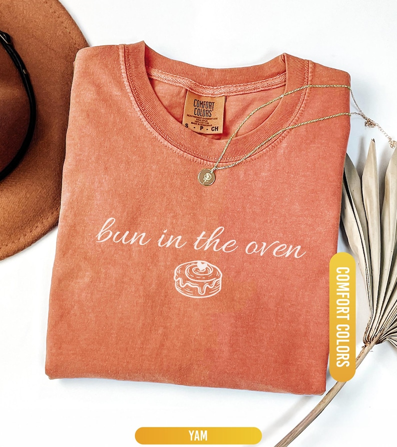 May include: A coral-colored t-shirt with the phrase "bun in the oven" printed in white, along with a graphic of a cinnamon roll. The shirt has a crew neck and a small tag that says "Comfort Colors". A gold necklace and a brown hat are also visible.