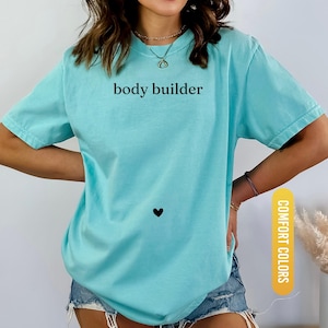 Pu&ograve; includere: Una maglietta azzurra con le parole "body builder" stampate in nero. Un piccolo cuore nero &egrave; sotto il testo. La maglietta &egrave; etichettata "Comfort Colors" e "Chalky Mint".