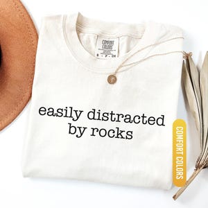 May include: Ivory-colored t-shirt with the phrase "easily distracted by rocks" in black lettering. A gold necklace with a pendant rests on the shirt. The shirt is labeled "Comfort Colors". A brown hat and dried leaves are in the background.
