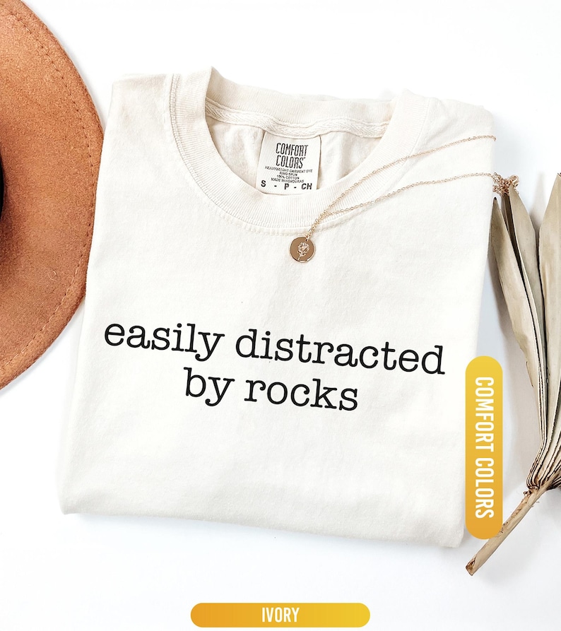 May include: Ivory-colored t-shirt with the phrase "easily distracted by rocks" in black lettering. A gold necklace with a pendant rests on the shirt. The shirt is labeled "Comfort Colors". A brown hat and dried leaves are in the background.