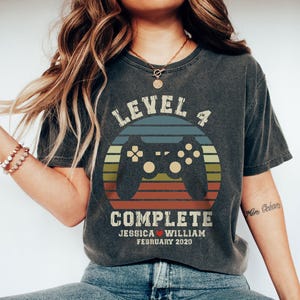 May include: Dark gray t-shirt with a retro video game controller design. The shirt features the text "LEVEL 4 COMPLETE" above the names Jessica and William, with the date February 2020. The design includes a circular graphic with a controller.