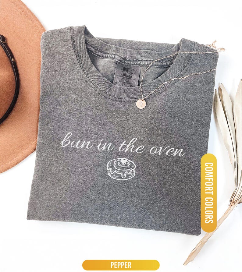 May include: A gray Comfort Colors t-shirt with the phrase "bun in the oven" and a drawing of a cinnamon roll. A gold necklace with a round pendant is on the shirt. The shirt is folded on a white surface.