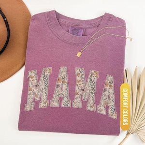 Op de afbeelding: Bes-kleurig T-shirt met het woord "MAMA" in bloemenprint. Het shirt heeft een ronde hals en een label met de tekst "COMFORT COLORS". Een gouden ketting met een rechthoekige hanger ligt op het shirt. Het woord "BERRY" staat op een geel label.