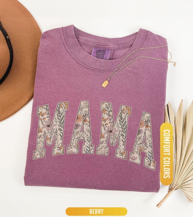 Op de afbeelding: Bes-kleurig T-shirt met het woord "MAMA" in bloemenprint. Het shirt heeft een ronde hals en een label met de tekst "COMFORT COLORS". Een gouden ketting met een rechthoekige hanger ligt op het shirt. Het woord "BERRY" staat op een geel label.