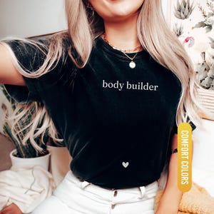 Pu&ograve; includere: T-shirt nero con la scritta bianca "body builder" e un piccolo cuore bianco. La maglietta &egrave; etichettata "Comfort Colors" e "Black". La modella indossa una collana d'oro.