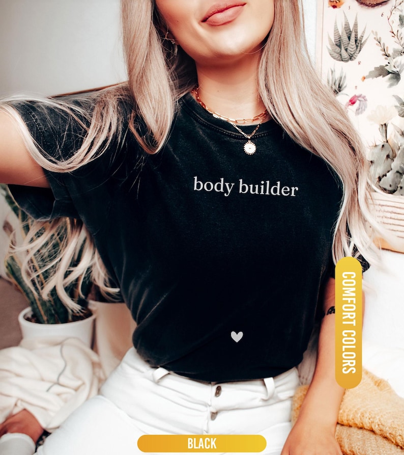 Pu&ograve; includere: T-shirt nero con la scritta bianca "body builder" e un piccolo cuore bianco. La maglietta &egrave; etichettata "Comfort Colors" e "Black". La modella indossa una collana d'oro.