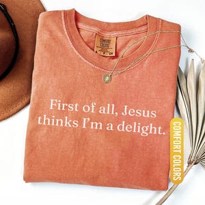 May include: A coral-colored t-shirt with the text "First of all, Jesus thinks I'm a delight." A gold necklace with a pendant rests on the shirt. The shirt has a Comfort Colors tag and a "YAM" label.