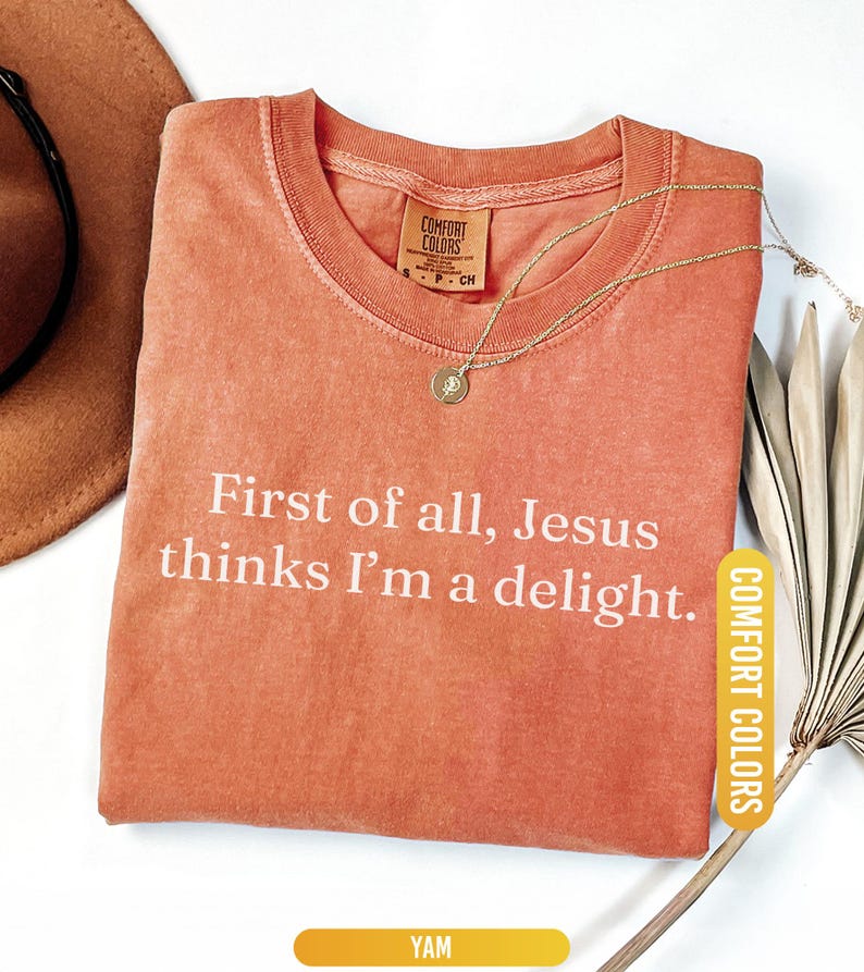 May include: A coral-colored t-shirt with the text "First of all, Jesus thinks I'm a delight." A gold necklace with a pendant rests on the shirt. The shirt has a Comfort Colors tag and a "YAM" label.