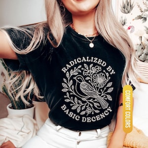 May include: Black t-shirt with a white graphic of a bird, flowers, and the text "RADICALIZED BY BASIC DECENCY". The shirt is labeled "BLACK" and "COMFORT COLORS".