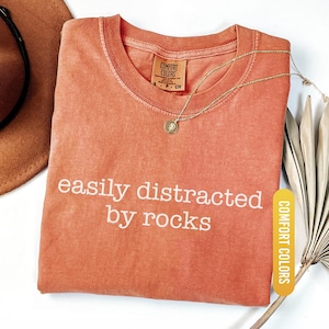 May include: A coral-colored t-shirt with the phrase "easily distracted by rocks" in white lettering. A gold necklace with a pendant rests on the shirt. The shirt has a Comfort Colors tag and a "YAM" label.