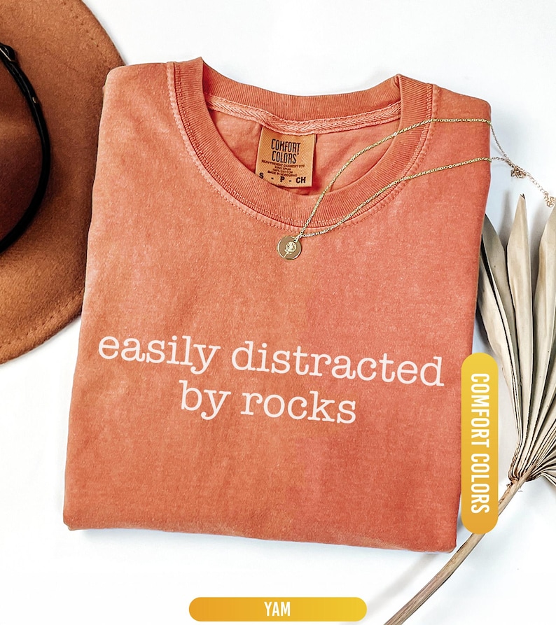 May include: A coral-colored t-shirt with the phrase "easily distracted by rocks" in white lettering. A gold necklace with a pendant rests on the shirt. The shirt has a Comfort Colors tag and a "YAM" label.