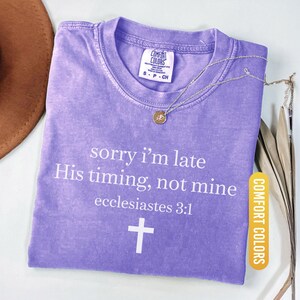 May include: A violet Comfort Colors t-shirt with the text "sorry i'm late His timing, not mine ecclesiastes 3:1" and a white cross. A silver necklace with a pendant is on the shirt.