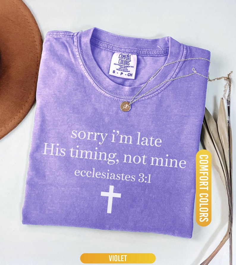 May include: A violet Comfort Colors t-shirt with the text "sorry i'm late His timing, not mine ecclesiastes 3:1" and a white cross. A silver necklace with a pendant is on the shirt.