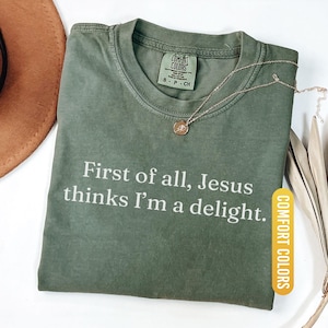 May include: A moss green Comfort Colors t-shirt with the text "First of all, Jesus thinks I'm a delight." A gold-colored necklace with a pendant is draped on the shirt. The shirt is folded on a white surface.