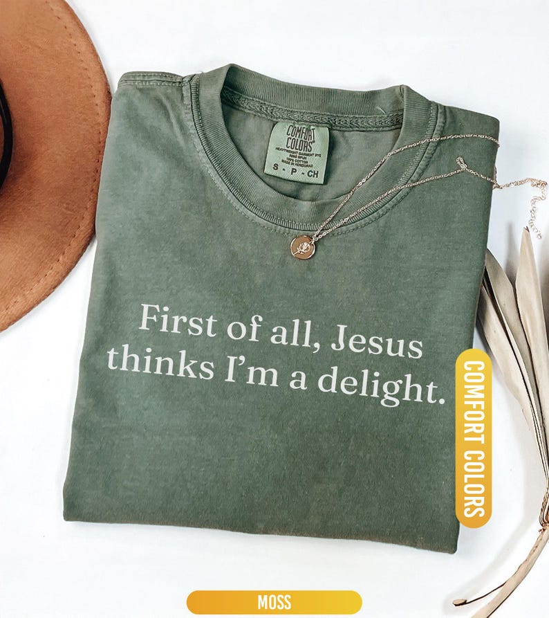 May include: A moss green Comfort Colors t-shirt with the text "First of all, Jesus thinks I'm a delight." A gold-colored necklace with a pendant is draped on the shirt. The shirt is folded on a white surface.