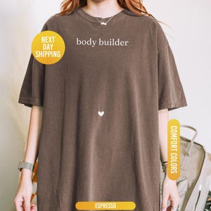 Pu&ograve; includere: T-shirt color espresso con la scritta bianca "body builder". La maglietta ha un piccolo cuore bianco e l'etichetta "Comfort Colors". Un cerchio giallo indica "Next Day Shipping".