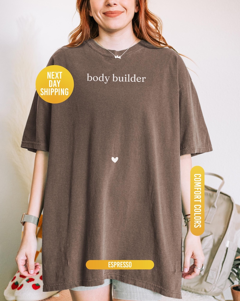 Pu&ograve; includere: T-shirt color espresso con la scritta bianca "body builder". La maglietta ha un piccolo cuore bianco e l'etichetta "Comfort Colors". Un cerchio giallo indica "Next Day Shipping".