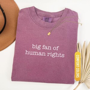 May include: A berry-colored t-shirt with the text "big fan of human rights" in white. A gold necklace with a rectangular pendant rests on the shirt. The shirt is labeled "Comfort Colors" and "Berry".