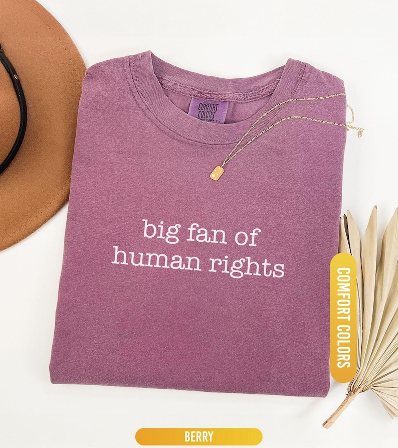 May include: A berry-colored t-shirt with the text "big fan of human rights" in white. A gold necklace with a rectangular pendant rests on the shirt. The shirt is labeled "Comfort Colors" and "Berry".