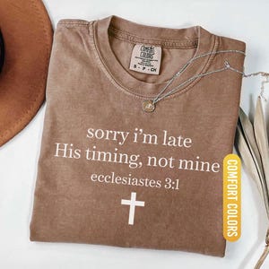 May include: Espresso-colored Comfort Colors t-shirt with the text "sorry I'm late His timing, not mine ecclesiastes 3:1" and a white cross. A silver necklace with a pendant is on the shirt.