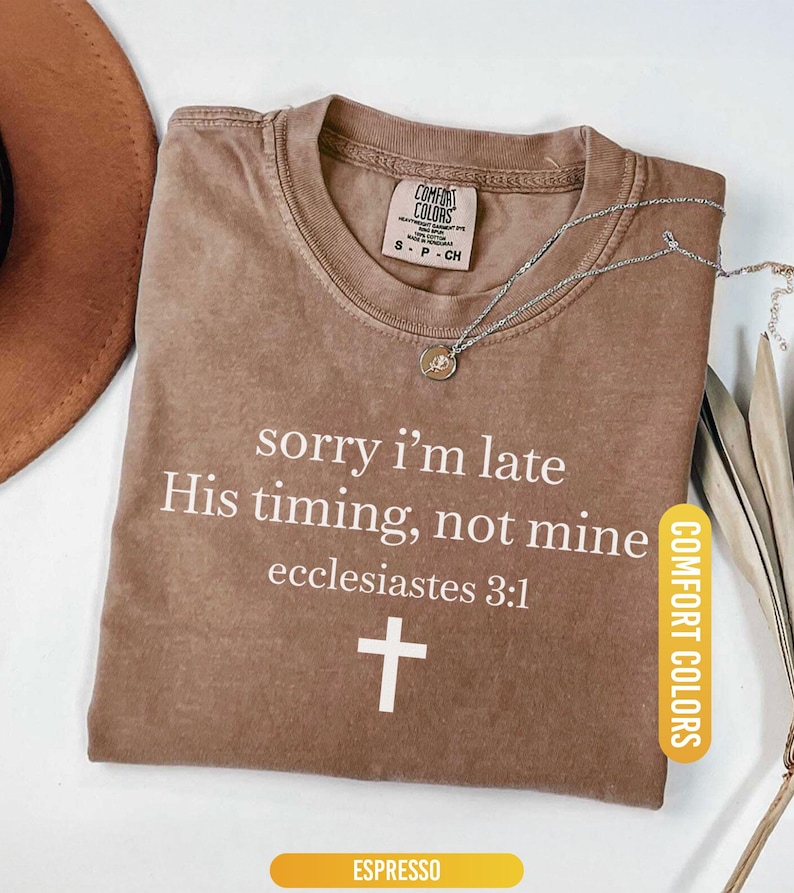 May include: Espresso-colored Comfort Colors t-shirt with the text "sorry I'm late His timing, not mine ecclesiastes 3:1" and a white cross. A silver necklace with a pendant is on the shirt.