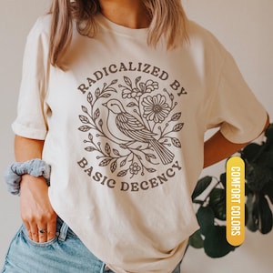 May include: Ivory-colored t-shirt with a circular graphic featuring a bird, flowers, and the text "RADICALIZED BY BASIC DECENCY." The shirt has short sleeves and a crew neck. The words "COMFORT COLORS" and "IVORY" are also visible.