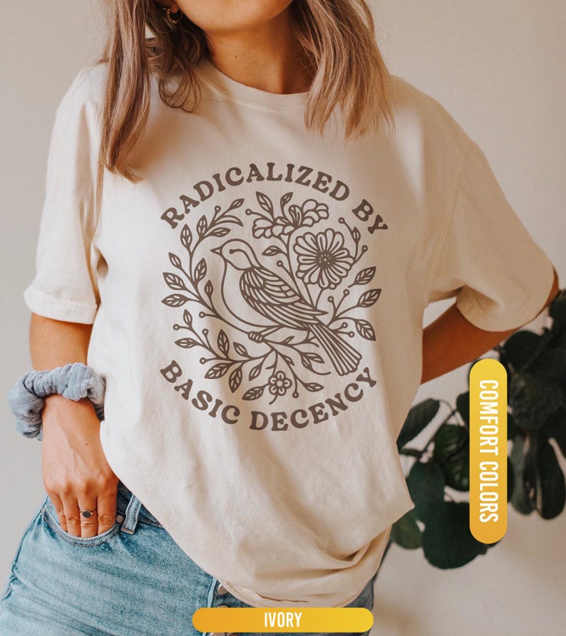 May include: Ivory-colored t-shirt with a circular graphic featuring a bird, flowers, and the text "RADICALIZED BY BASIC DECENCY." The shirt has short sleeves and a crew neck. The words "COMFORT COLORS" and "IVORY" are also visible.
