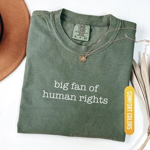May include: A moss green Comfort Colors t-shirt with the text "big fan of human rights" in white. A gold necklace with a small pendant rests on the shirt. The shirt is folded and displayed on a white surface.