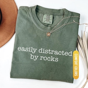 May include: A moss green Comfort Colors t-shirt with the text "easily distracted by rocks" in white. A gold necklace with a small pendant rests on the shirt. A brown hat is partially visible in the upper left corner.
