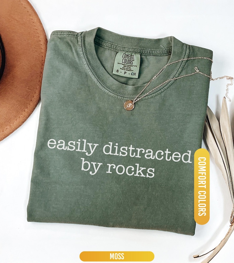 May include: A moss green Comfort Colors t-shirt with the text "easily distracted by rocks" in white. A gold necklace with a small pendant rests on the shirt. A brown hat is partially visible in the upper left corner.