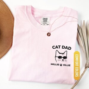 May include: A light pink t-shirt with the words "CAT DAD" and a cat wearing sunglasses. The names "Millie" and "Ollie" are printed below the cat image. The shirt has a "Comfort Colors" tag and a "Blossom" color label.
