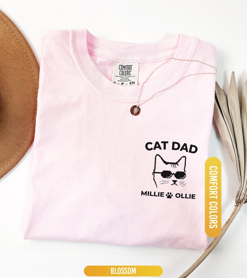 May include: A light pink t-shirt with the words "CAT DAD" and a cat wearing sunglasses. The names "Millie" and "Ollie" are printed below the cat image. The shirt has a "Comfort Colors" tag and a "Blossom" color label.