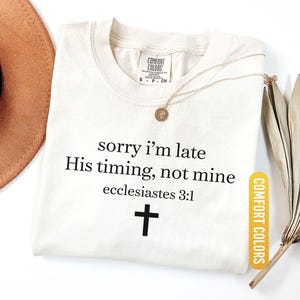 May include: Ivory-colored t-shirt with the text "sorry I'm late, His timing, not mine, ecclesiastes 3:1" and a black cross. A gold necklace with a pendant rests on the shirt. The shirt is labeled "Comfort Colors".