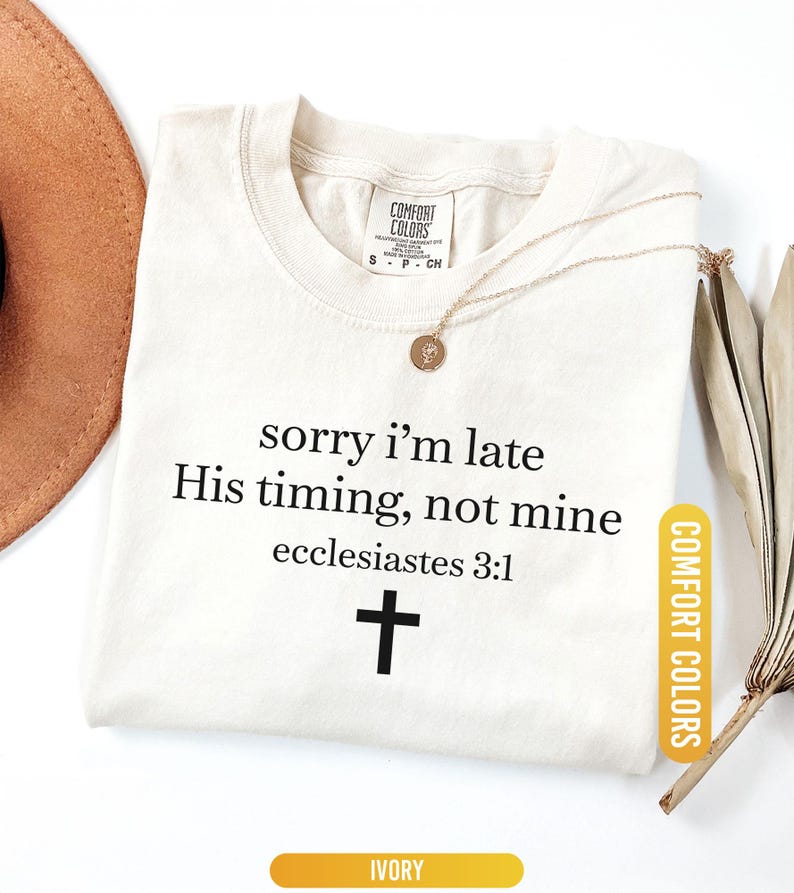 May include: Ivory-colored t-shirt with the text "sorry I'm late, His timing, not mine, ecclesiastes 3:1" and a black cross. A gold necklace with a pendant rests on the shirt. The shirt is labeled "Comfort Colors".