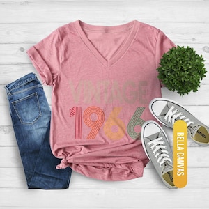 Pu&ograve; includere: Una maglietta con scollo a V color malva melange con la scritta "VINTAGE 1966" in colori retr&ograve;. La maglietta &egrave; abbinata a jeans blu e scarpe da ginnastica grigie. C'&egrave; anche una piccola pianta verde nell'immagine. La maglietta &egrave; di Bella Canvas.