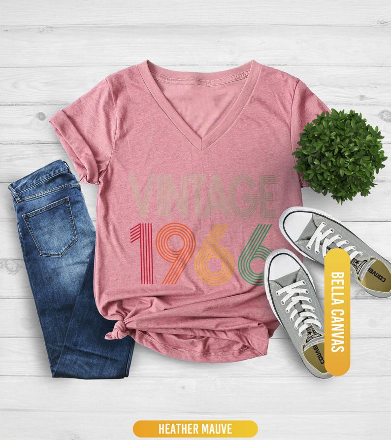 Pu&ograve; includere: Una maglietta con scollo a V color malva melange con la scritta "VINTAGE 1966" in colori retr&ograve;. La maglietta &egrave; abbinata a jeans blu e scarpe da ginnastica grigie. C'&egrave; anche una piccola pianta verde nell'immagine. La maglietta &egrave; di Bella Canvas.