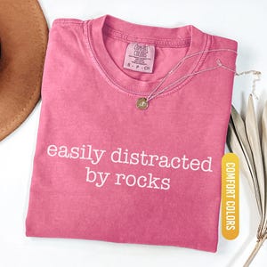 May include: A pink t-shirt with the text "easily distracted by rocks" in white. A silver necklace with a pendant rests on the shirt. The shirt is labeled "Comfort Colors" and is a size small.
