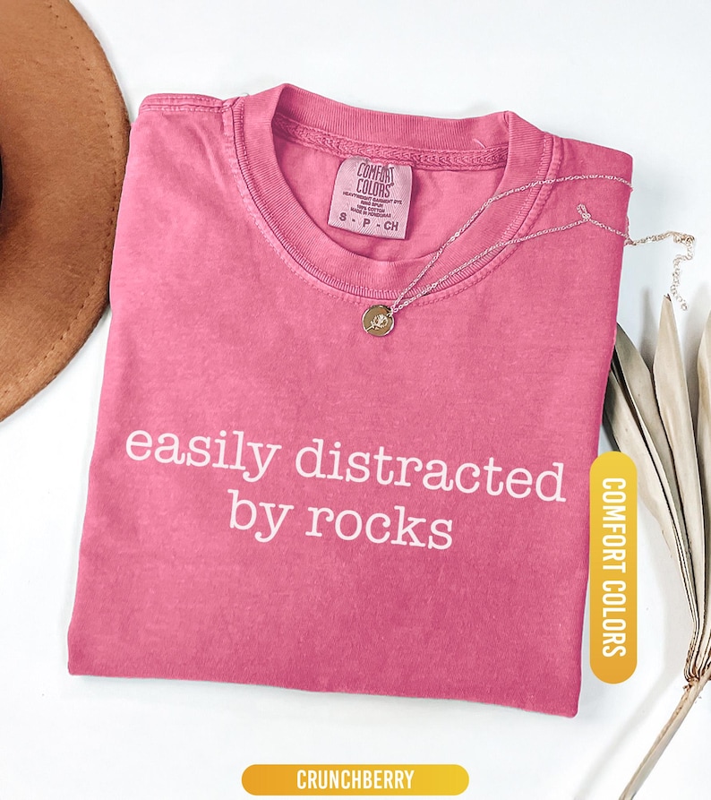 May include: A pink t-shirt with the text "easily distracted by rocks" in white. A silver necklace with a pendant rests on the shirt. The shirt is labeled "Comfort Colors" and is a size small.